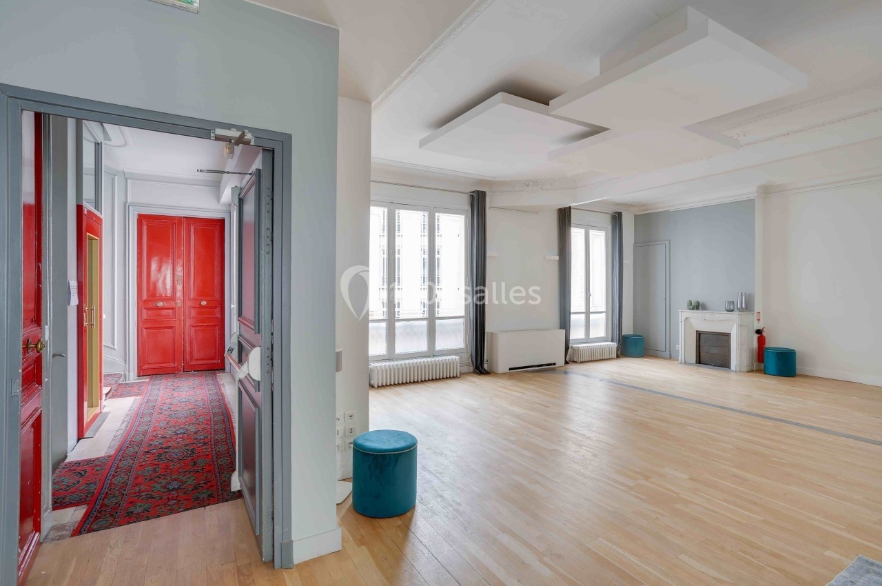 Salle lumineuse avec parquet clair, grandes fenêtres, murs blancs, poufs bleus et une porte rouge visible au fond.