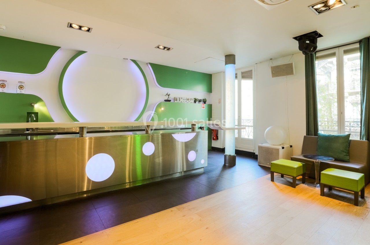 Intérieur moderne avec comptoir en métal, murs verts et blancs, éclairage design et coin salon avec fauteuils.