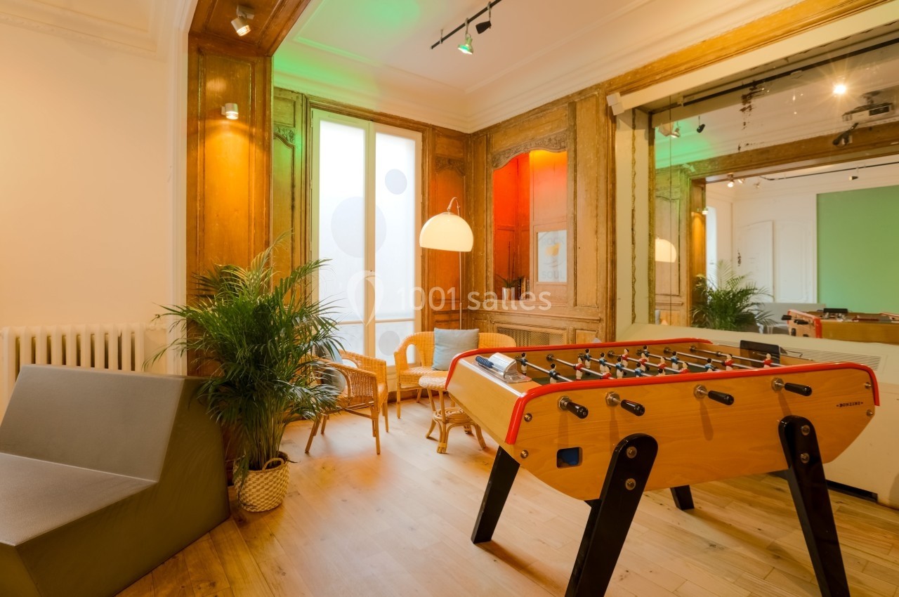 Salle lumineuse avec baby-foot, fauteuils en rotin, plantes et grandes fenêtres entourées de boiseries.