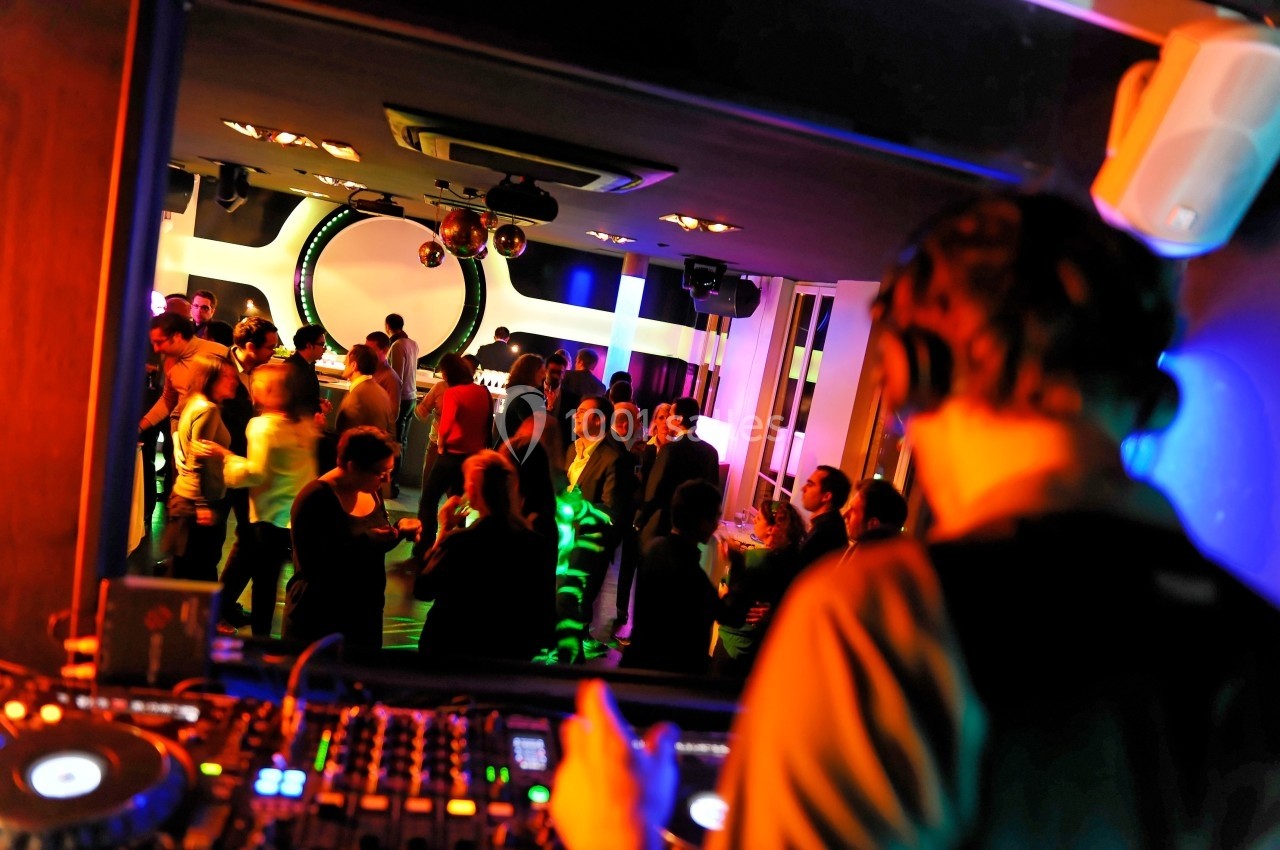 Un DJ mixe de la musique dans une salle animée où des personnes dansent et discutent sous des lumières colorées.