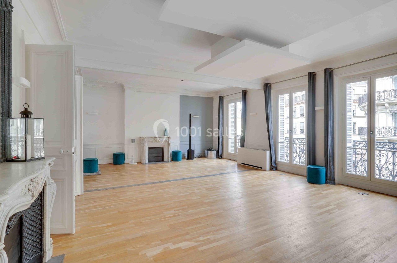 L'Appart Lafayette – espace lumineux pour cocktail et réception Salle lumineuse avec parquet clair, cheminée décorative, grandes fenêtres et rideaux gris donnant sur un balcon en fer forgé.