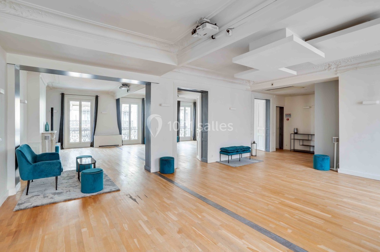 L'Appart Lafayette – espace modulable pour réception privée Salle lumineuse avec parquet clair, fauteuils bleus, grandes fenêtres et éléments de décoration modernes.