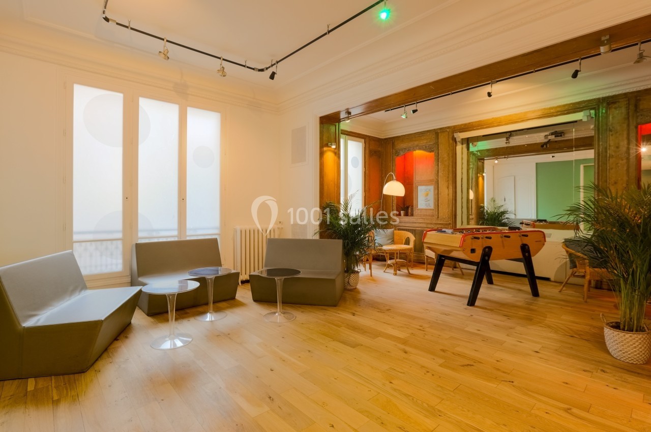 Salon Lafayette – espace modulable pour cocktail et soirée Salle lumineuse avec parquet, canapés gris, baby-foot, plantes et grandes fenêtres laissant entrer la lumière naturelle.