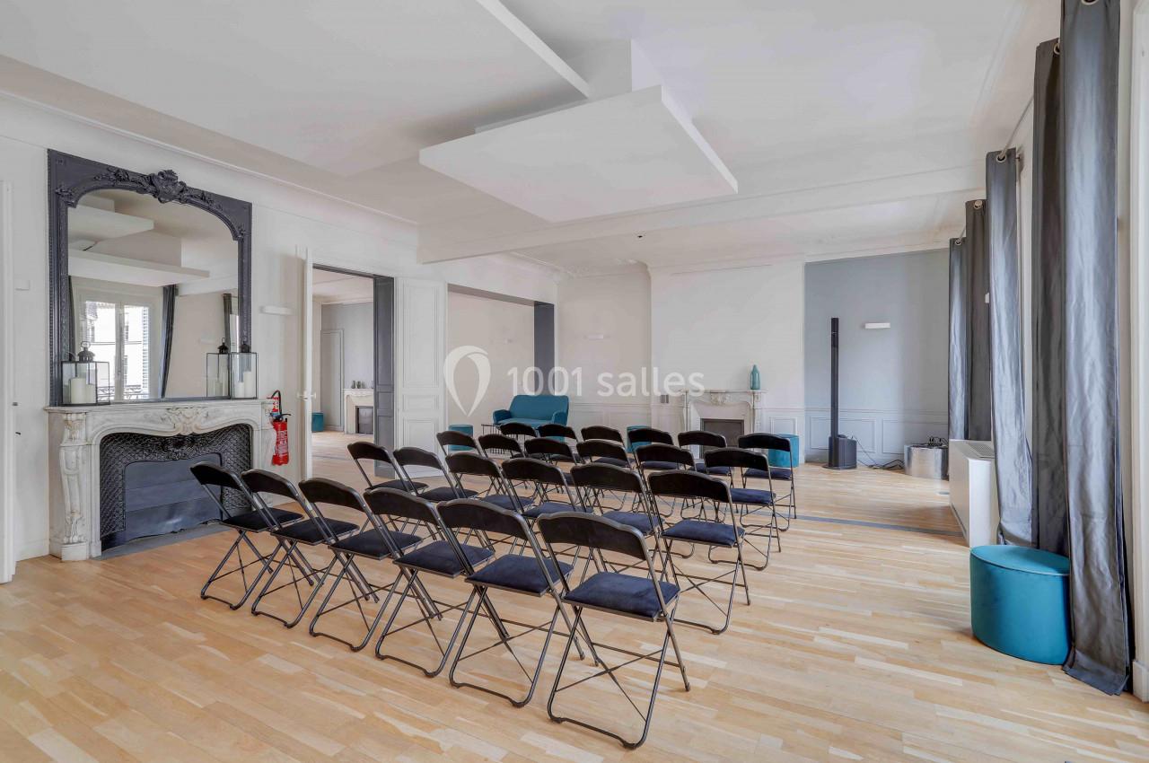 Salle lumineuse avec parquet, chaises pliantes alignées, cheminée décorative et grandes fenêtres avec rideaux.