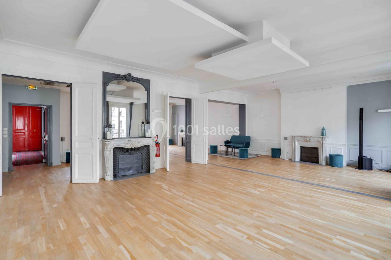 Salle lumineuse avec parquet clair, cheminée décorative, murs blancs et une porte rouge visible au fond.