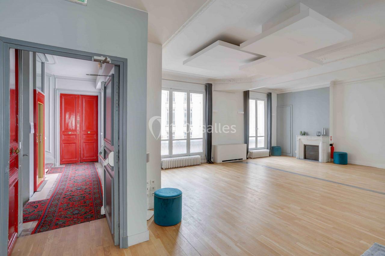 Salle lumineuse avec parquet clair, grandes fenêtres, plafonniers modernes et portes rouges visibles dans une autre pièce.