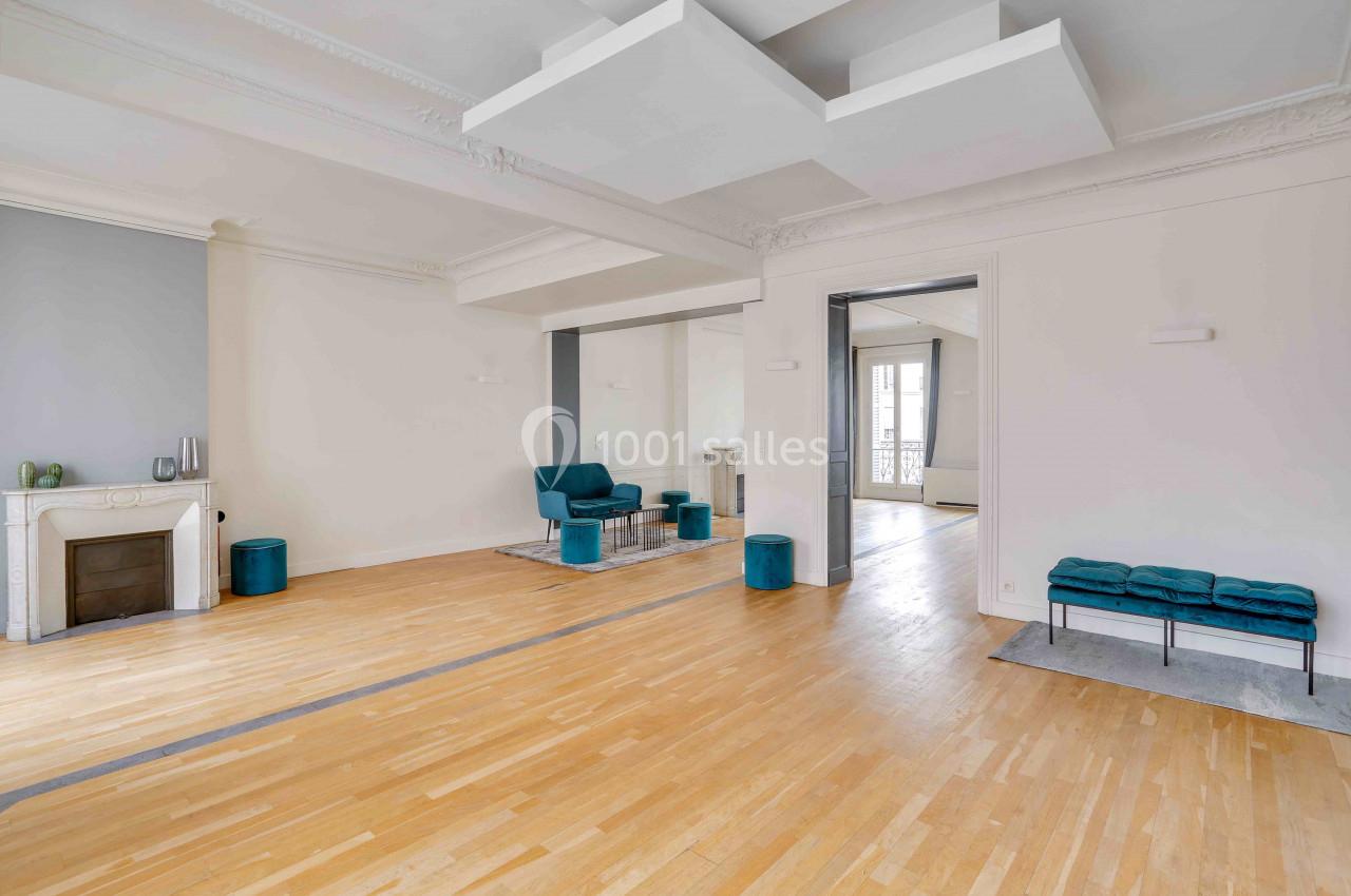 Salon lumineux avec parquet clair, cheminée décorative, murs blancs, fauteuils bleu-vert et moulures au plafond.