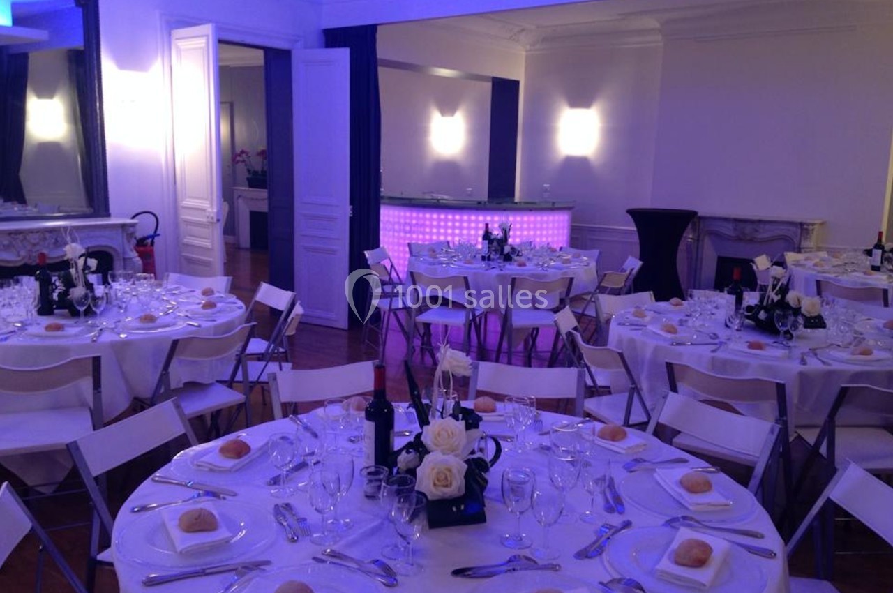 Salle de réception élégante avec tables rondes dressées, nappes blanches, couverts et éclairage tamisé violet.