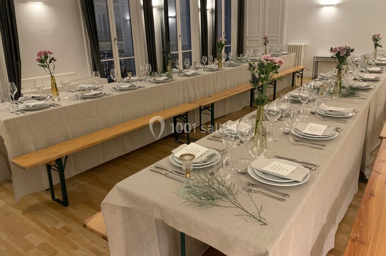 Tables longues dressées avec nappes beiges, vaisselle élégante, bancs en bois et décorations florales dans une salle…