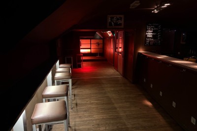 Miniature Location salle Lyon 1 (Rhône) - Le Temps des Copains #9 Salle sombre avec un éclairage rouge au fond, tabourets alignés à gauche et un comptoir à droite.