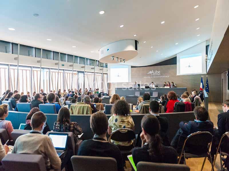 Salle de conférence avec des participants assis, regardant une présentation projetée sur deux écrans.