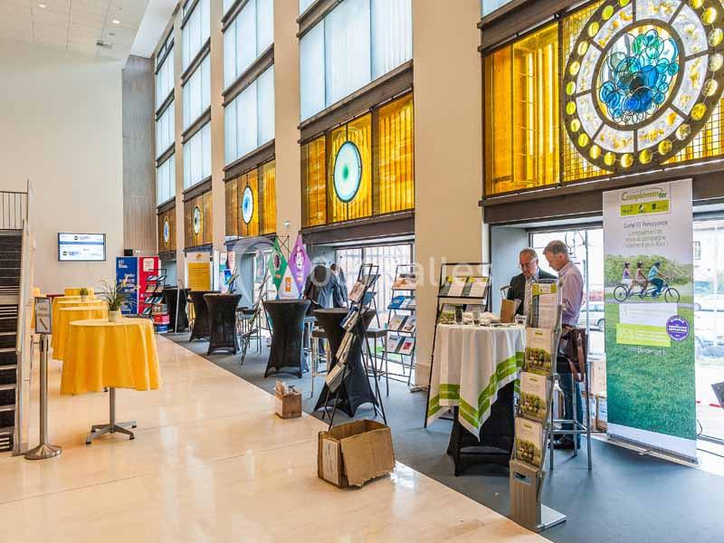 Salle d'exposition lumineuse avec des stands d'information, des affiches et des participants en discussion.