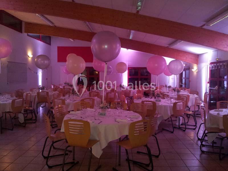 Salle de réception décorée avec des ballons roses et tables rondes dressées pour un événement festif.