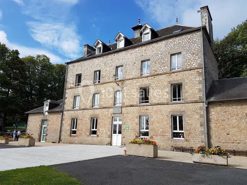 Bâtiment en pierre à trois étages avec fenêtres blanches, entouré de végétation et d'un espace pavé.