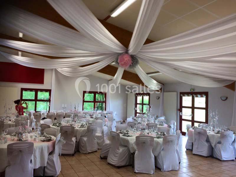 Salle de réception décorée avec des drapés blancs au plafond, tables rondes dressées et chaises couvertes de housses…