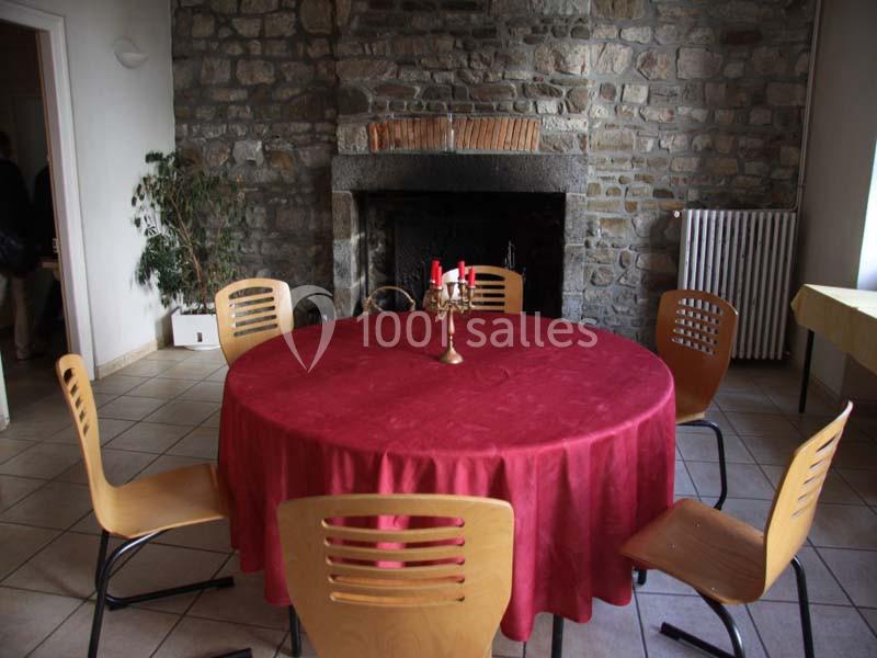Table ronde recouverte d'une nappe rouge, entourée de chaises en bois, devant une cheminée en pierre.