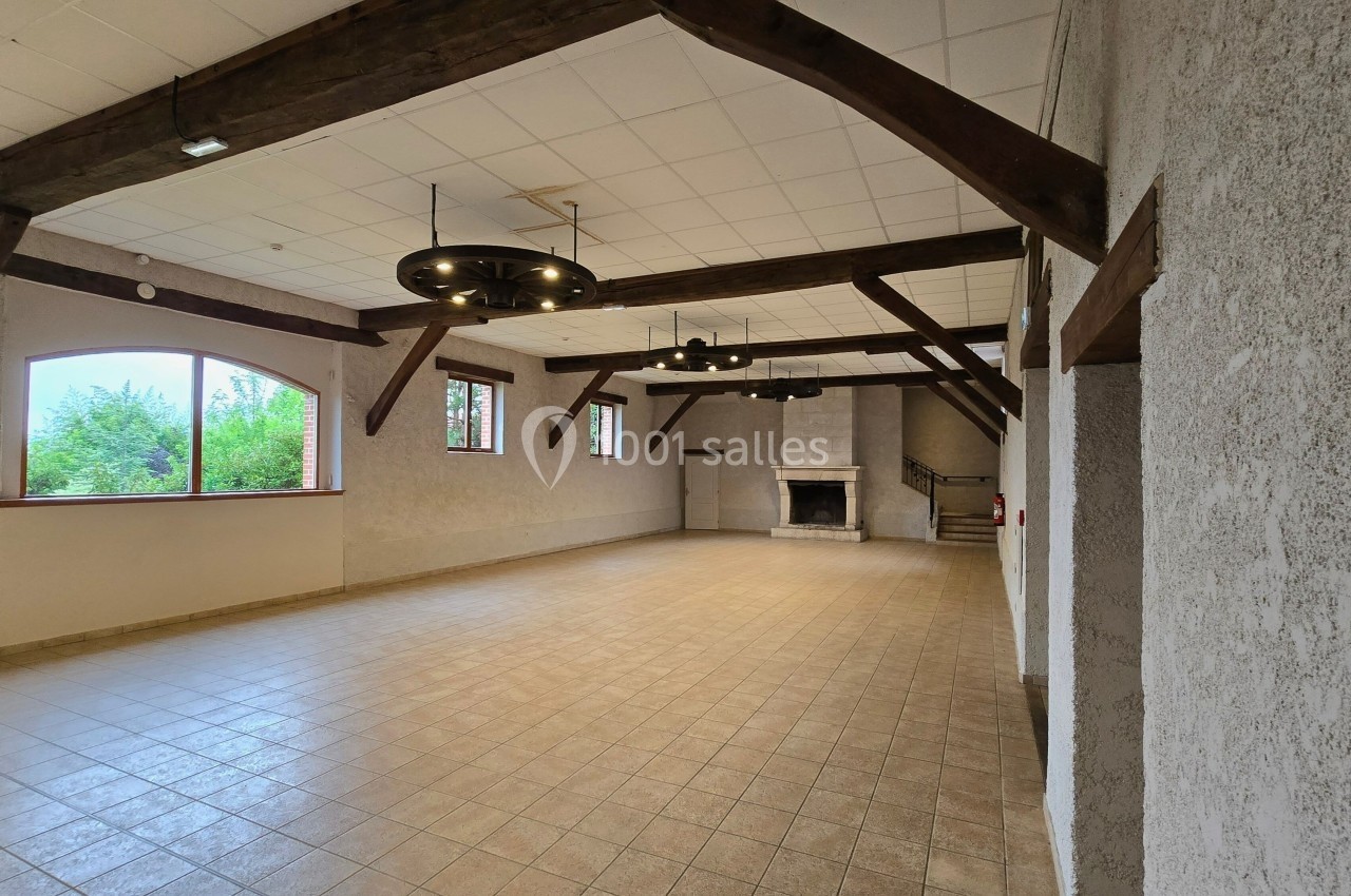 Salle spacieuse avec poutres apparentes, cheminée en pierre, grandes fenêtres et carrelage beige.