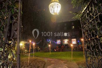 Jardin avec pelouse, tables et chaises rouges, scène en bois, entouré d'arbres et d'une maison à colombages.