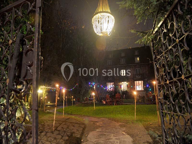 Entrée d'un jardin illuminé par des torches et un lustre suspendu, avec une maison en arrière-plan de nuit.