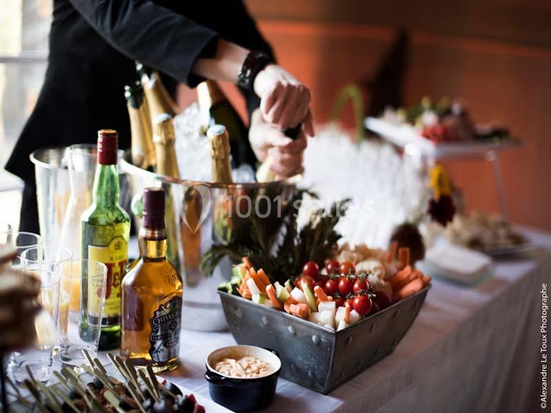Un serveur ouvre une bouteille près d'une table garnie de boissons, légumes, et apéritifs variés.
