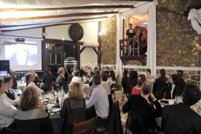 Jardin avec pelouse, tables et chaises rouges, scène en bois, entouré d'arbres et d'une maison à colombages.