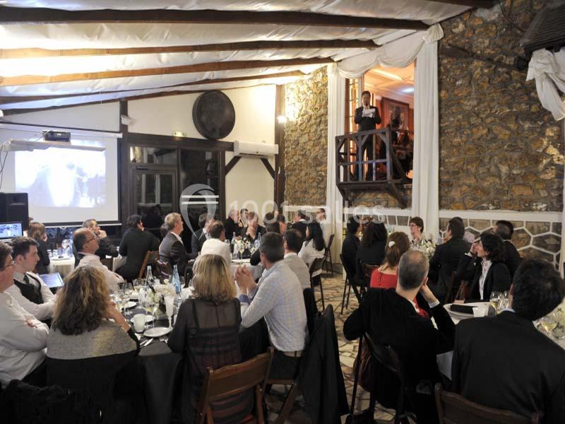 Des personnes assises à des tables lors d'un événement en intérieur, écoutant un orateur debout sur une mezzanine.
