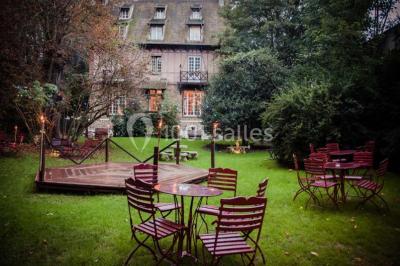 Jardin avec pelouse, tables et chaises rouges, scène en bois, entouré d'arbres et d'une maison à colombages.