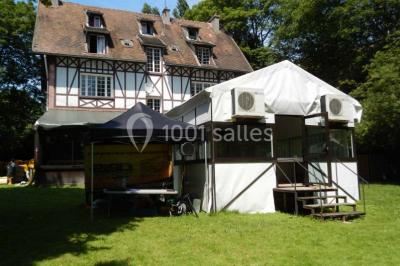 Jardin avec pelouse, tables et chaises rouges, scène en bois, entouré d'arbres et d'une maison à colombages.