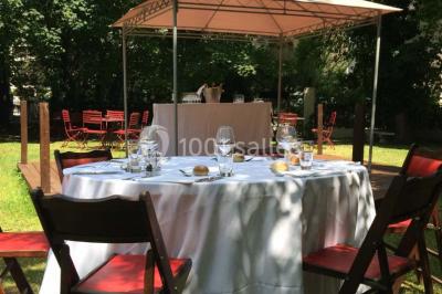 Jardin avec pelouse, tables et chaises rouges, scène en bois, entouré d'arbres et d'une maison à colombages.