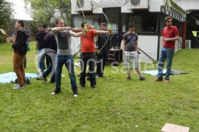 Jardin avec pelouse, tables et chaises rouges, scène en bois, entouré d'arbres et d'une maison à colombages.