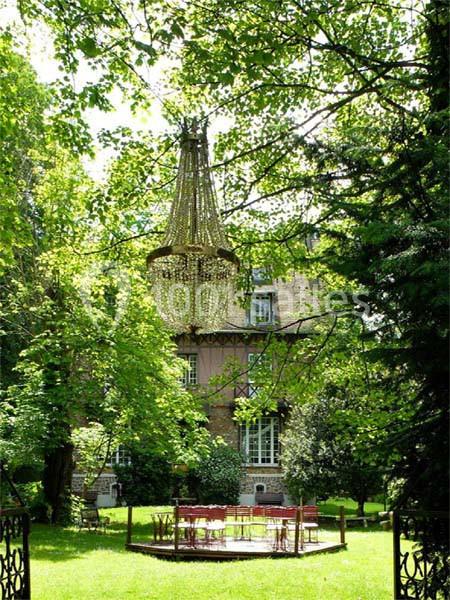 Jardin verdoyant avec un grand lustre suspendu aux arbres, des tables et chaises rouges disposées sur une pelouse.