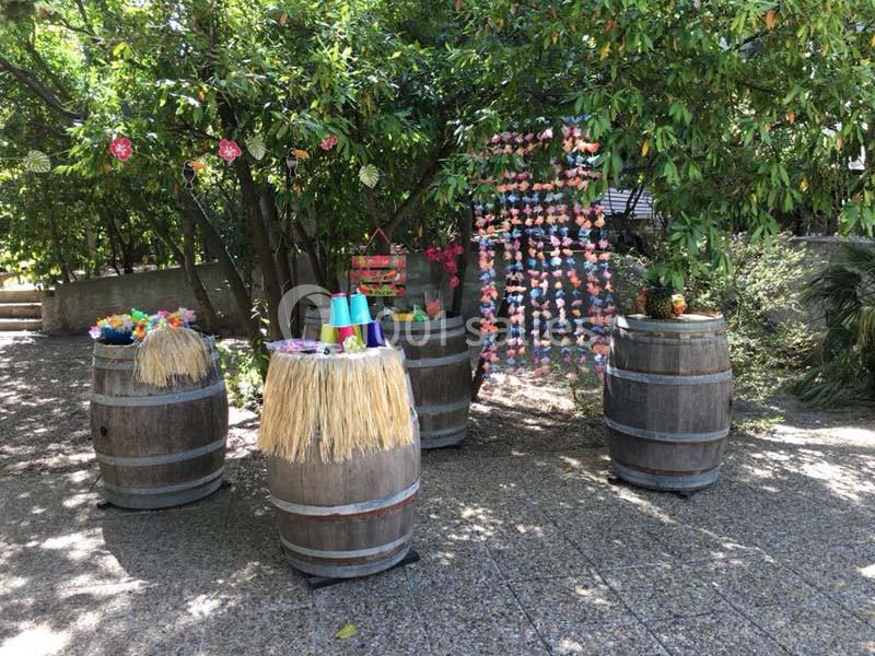 Décoration festive en extérieur avec tonneaux, guirlandes colorées et accessoires sur un sol pavé sous des arbres.