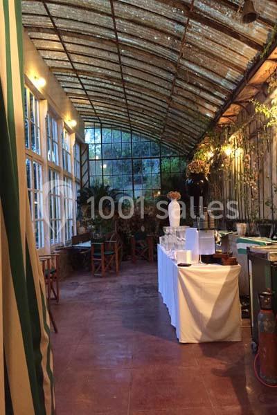 Salle lumineuse avec verrière, table dressée pour un buffet, plantes et chaises en bois en arrière-plan.