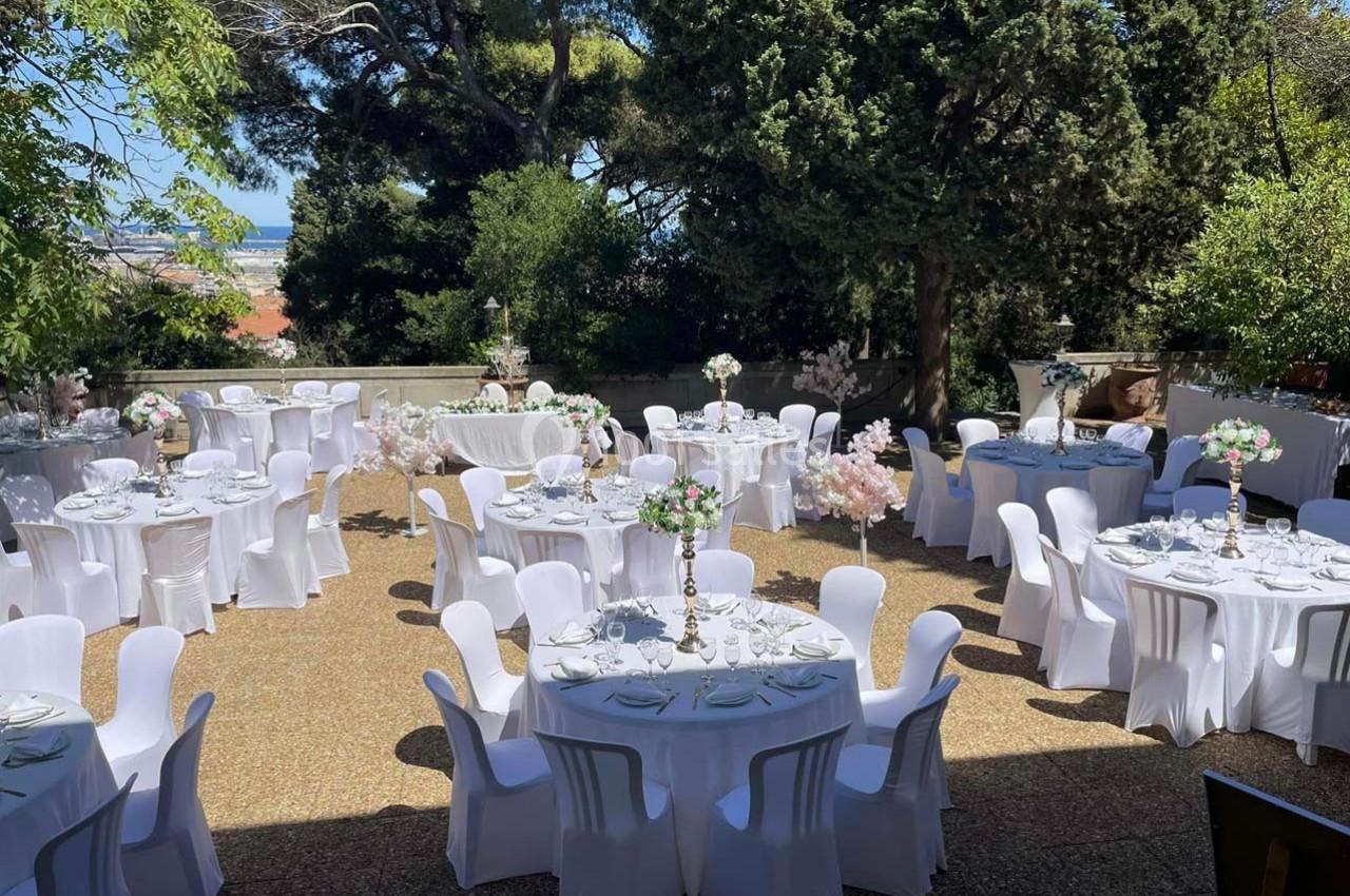 Tables rondes décorées pour un événement en plein air, entourées de verdure et disposées sur une terrasse ensoleillée.
