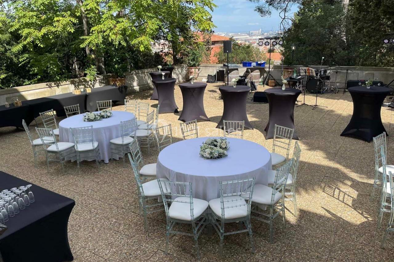 Terrasse aménagée pour un événement avec tables rondes dressées, chaises transparentes et tables hautes noires.