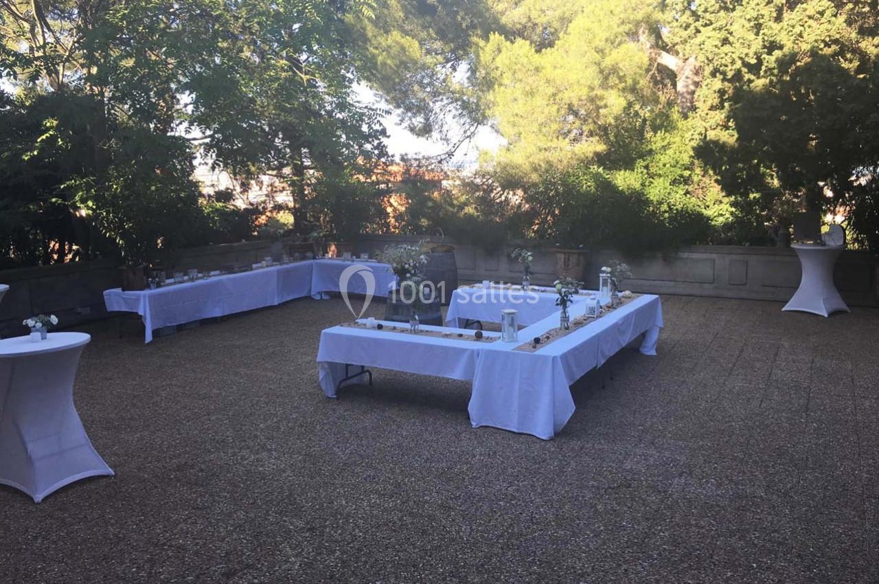 Tables rectangulaires dressées avec nappes blanches et décorations, disposées en extérieur dans un jardin ombragé.