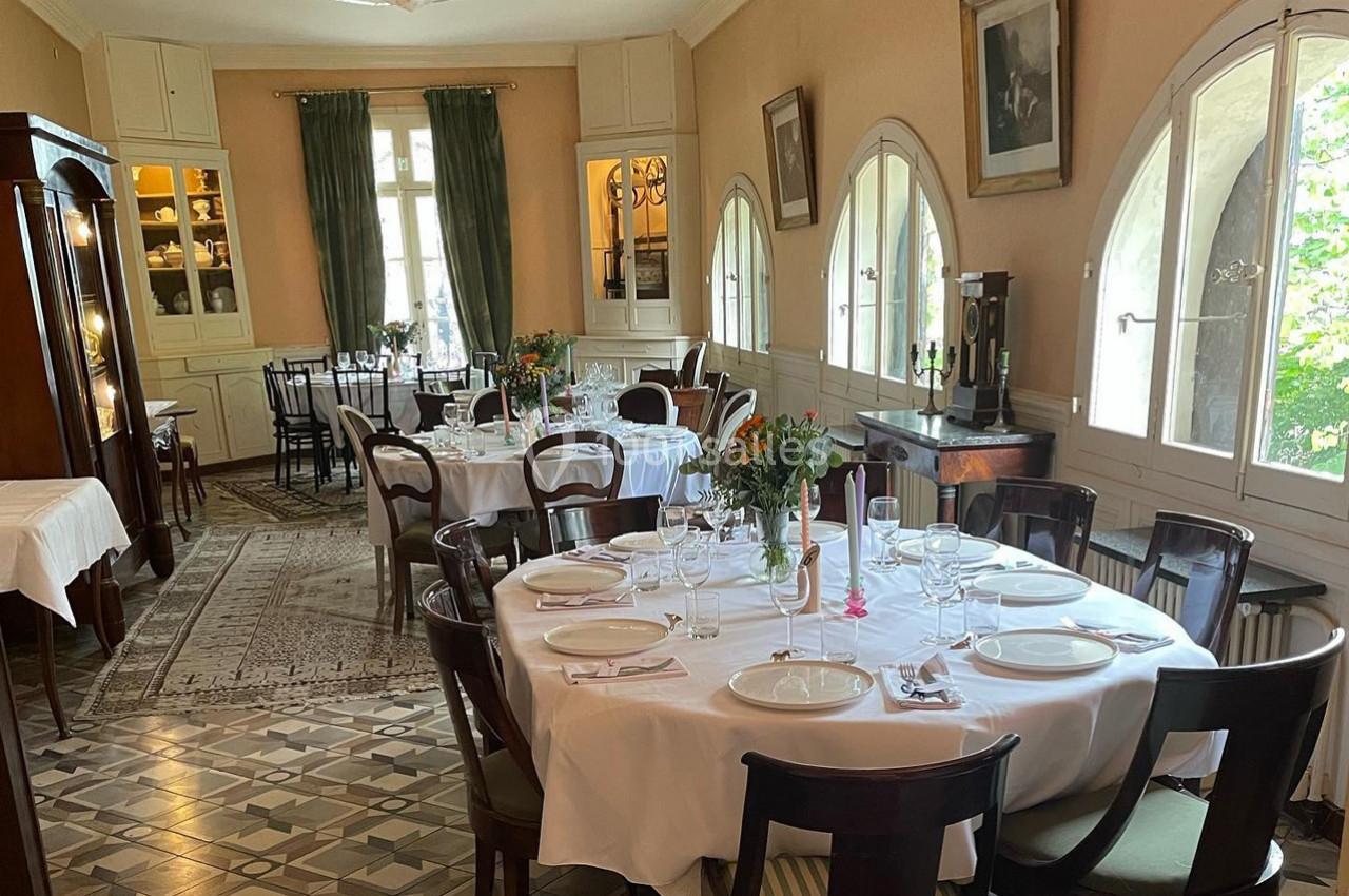 Salle à manger élégante avec tables rondes dressées, chaises en bois, grandes fenêtres et décoration classique.