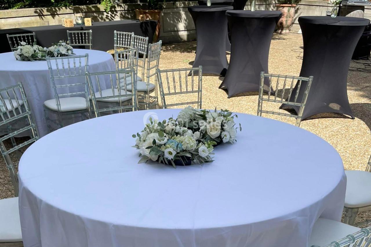 Table ronde décorée de fleurs blanches, entourée de chaises transparentes, dans un espace extérieur ensoleillé.