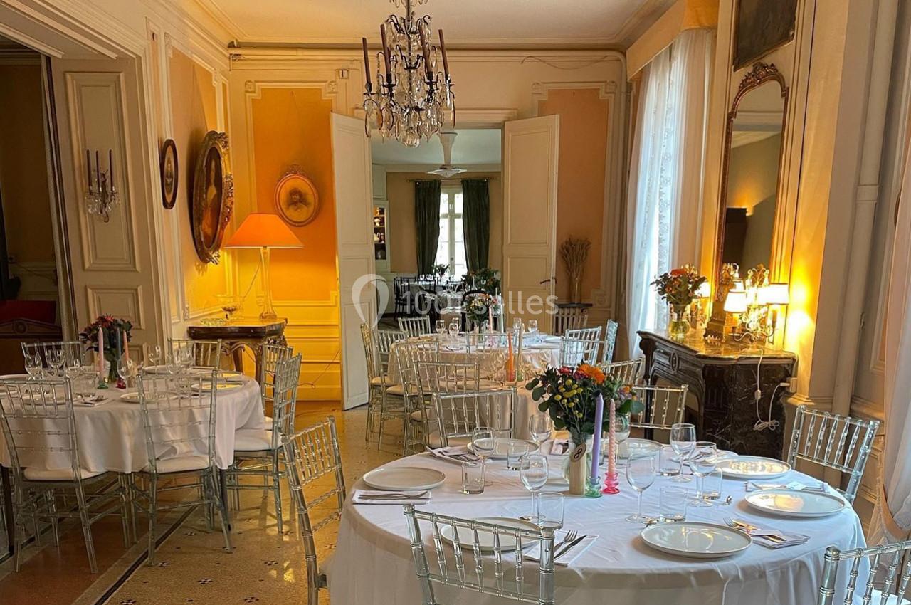 Salle élégante avec tables rondes dressées, chaises transparentes, lustre, miroirs et lumière naturelle.