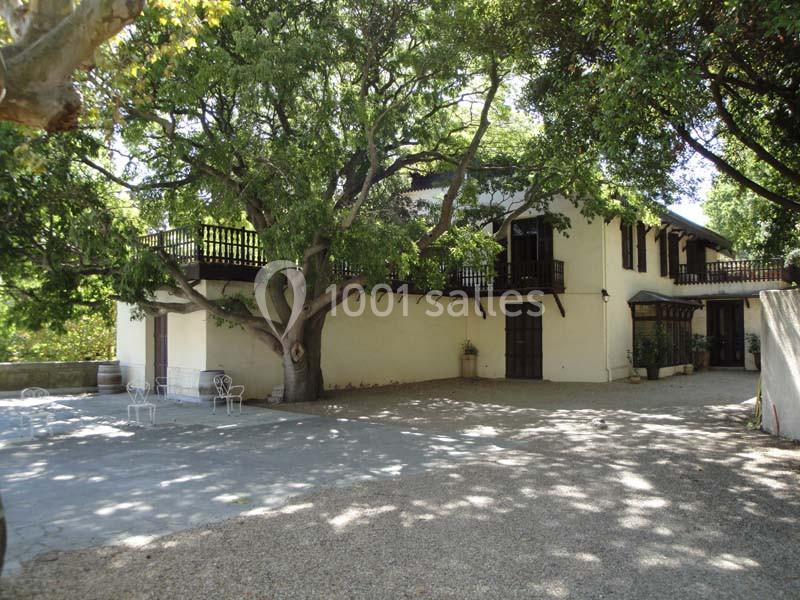 Grande maison blanche entourée d'arbres, avec balcons en bois et une cour ombragée par un grand arbre.