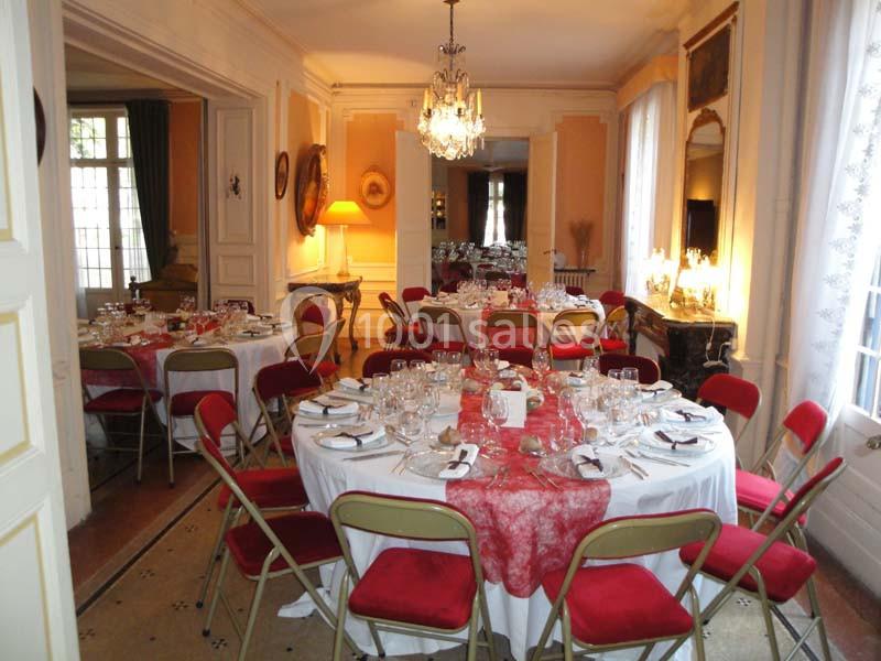 Salle élégante avec tables rondes dressées pour un repas, nappes blanches, chaises rouges et lustre au plafond.