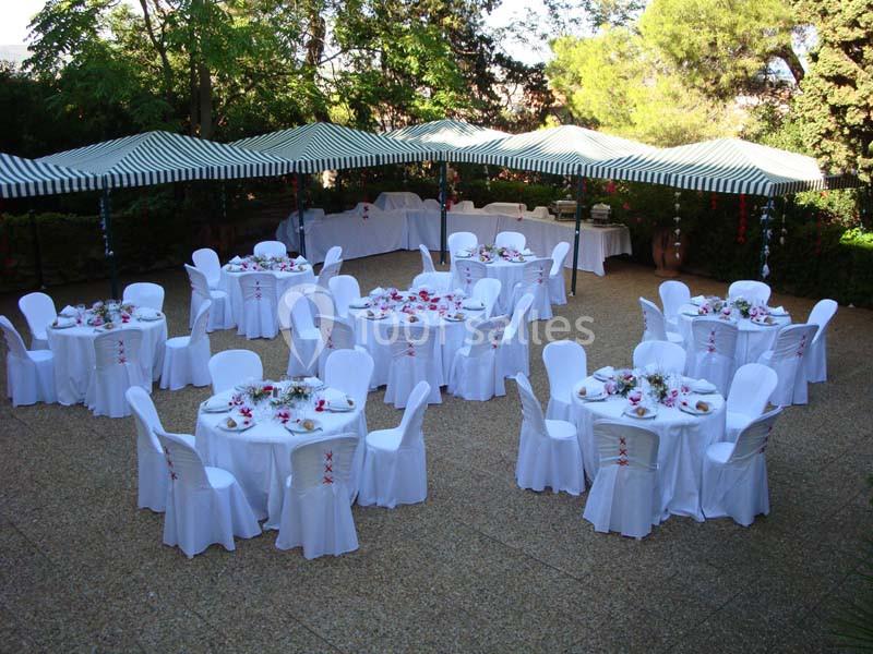 Tables rondes décorées avec nappes blanches et arrangements floraux, disposées sous des parasols rayés dans un jardin.