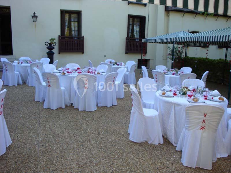 Tables rondes dressées avec nappes blanches et chaises décorées, disposées dans une cour extérieure pavée.