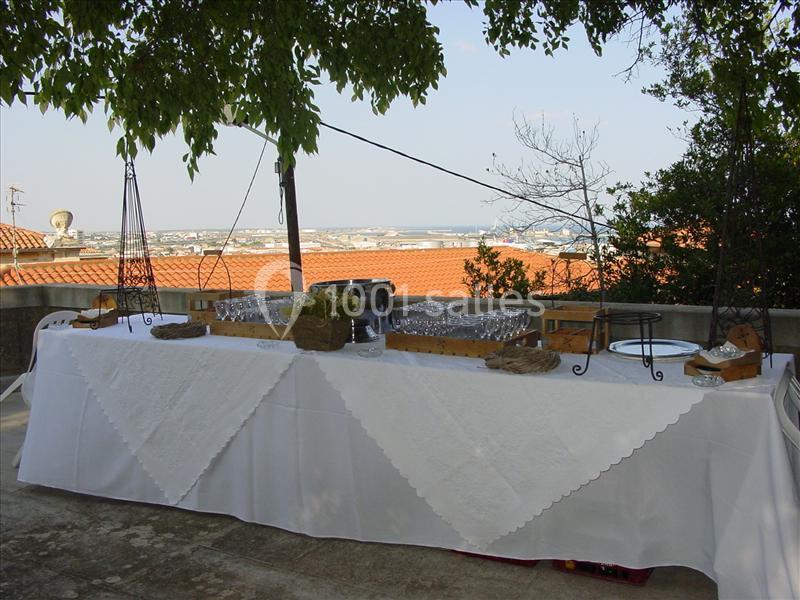 Table dressée en extérieur avec nappe blanche, verres, paniers et vue sur des toits et un paysage lointain.