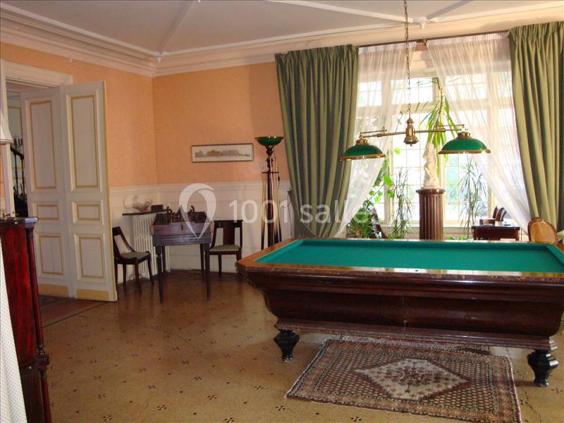 Salle avec un billard en bois, éclairé par des lampes suspendues, entouré de meubles anciens et de grandes fenêtres.