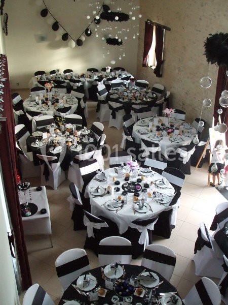 Salle décorée pour un événement avec des tables rondes dressées, nappes blanches et noires, et décorations suspendues.