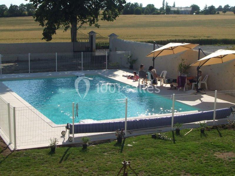 Piscine extérieure entourée d'une clôture, avec des parasols et des personnes assises sur des chaises près de l'eau.