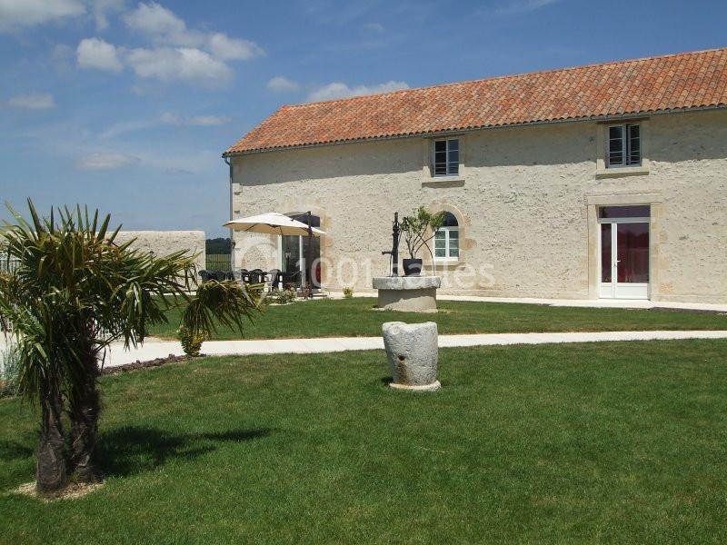 Façade d'une maison en pierre avec toit en tuiles, terrasse aménagée, puits ancien et pelouse arborée.