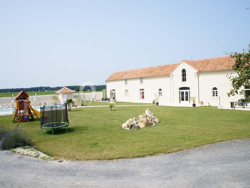 Grande maison en pierre avec toit en tuiles, jardin aménagé avec trampoline, aire de jeux et piscine en arrière-plan.