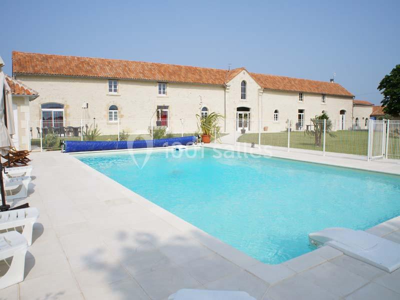 Piscine extérieure avec terrasse en pierre, entourée d'une clôture, devant un bâtiment en pierre à toit rouge.